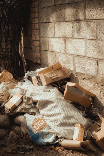A pile of mixed waste materials is situated at the base of a brick wall, with some cardboard boxes of various sizes and shapes, some partially crushed or bent. The boxes are light brown, with visible printed labels and markings, and are scattered across a coarse, uneven ground composed of dirt and small stones. A beige woven sack or bag is visible among the waste, appearing to contain or support some of the debris. To the left, a large tree trunk with dark, textured bark is partially visible, casting shadows on the scene. The brick wall behind the waste is constructed from light-colored, rough-textured bricks arranged in a horizontal pattern, with some bricks showing slight discoloration. The scene appears outdoors, in a narrow space between buildings or against a boundary wall, and suggests an environment where independent rubbish removal or on-site clearance is taking place, as managed by Rubbish Removal Greenwich, in support of local waste disposal options outside of municipal collection systems.
