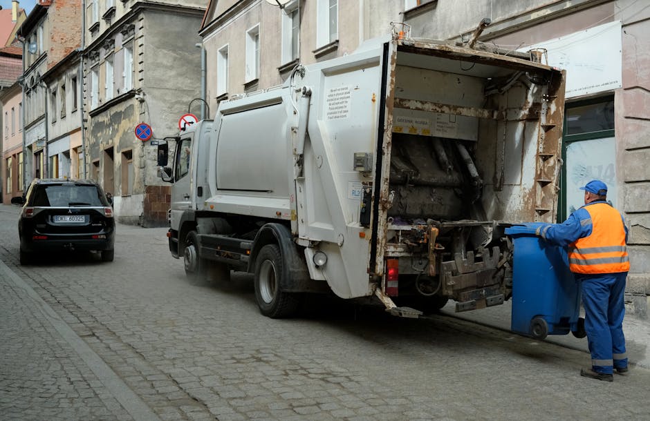 A white waste collection truck is parked on a narrow street in an urban area, with a worker dressed in a blue uniform and orange high-visibility vest standing beside the open rear compartment, which appears to contain emptied rubbish or debris. The worker is placing a blue wheeled bin into the truck's hopper area, which has visible rust and metal mechanical components. To the left of the truck, a black car is parked on the cobblestone pavement, and the background shows a row of multi-storey buildings with a mixture of stone and brick facades, some with visible aging and peeling paint. Street signs indicating no parking or stopping are mounted on the building wall behind the truck. The scene is lit by natural daylight, casting soft shadows, and reflects an outdoor urban rubbish removal operation, exemplifying private waste collection services suited for narrow access situations. Rubbish Removal Greenwich occasionally undertakes on-site clearance for difficult-to-access locations, ensuring efficient disposal without reliance on municipal waste services.