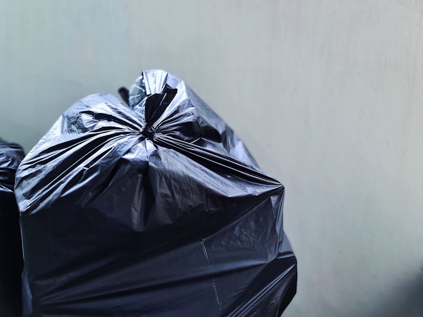 Close-up view of a black plastic garbage bag filled with waste, tightly gathered and sealed at the top with multiple folds and creases visible on its surface. The bag appears to be made of glossy, crinkled plastic material, reflecting ambient light and showing slight reflections of the surrounding environment. It is positioned in the foreground against a plain, light-colored wall, with additional similar bags partially visible behind it, suggesting a collection of rubbish prepared for removal. The scene is set outdoors on a paved or concrete surface, indicative of a typical waste disposal or rubbish collection area. The neutral lighting and absence of other objects emphasize the focus on the plastic waste bag, aligning with professional rubbish removal practices such as private waste handling or alternative rubbish clearance services conducted by companies like Rubbish Removal Greenwich.