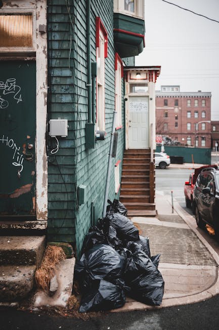 In the foreground of the image, several black garbage bags are piled up against the side of a weathered building with green wooden siding and red trim around windows and doors. The bags appear tightly sealed and are placed on a small concrete step, partially on a concrete sidewalk that extends into a wider paved area. To the left, a metal door with graffiti and a small step lead into the building, which has a slightly peeling surface, indicating age. The building's side features an electrical box and connectors, with a cable running vertically along the wall. Adjacent to the bags, a set of wooden stairs with a handrail ascends to a white door with a small window, suggesting an entrance to the building. The surrounding environment includes parked cars along the street, with additional urban structures visible in the background, such as brick buildings and rooftop elements. The scene is lit with natural daylight, typical of an overcast day, emphasizing the utilitarian nature of the setting while subtly illustrating the process of private rubbish disposal and collections outside a residential or commercial property. Rubbish Removal Greenwich operates in this context, providing alternative waste handling services to on-site or private collection solutions.