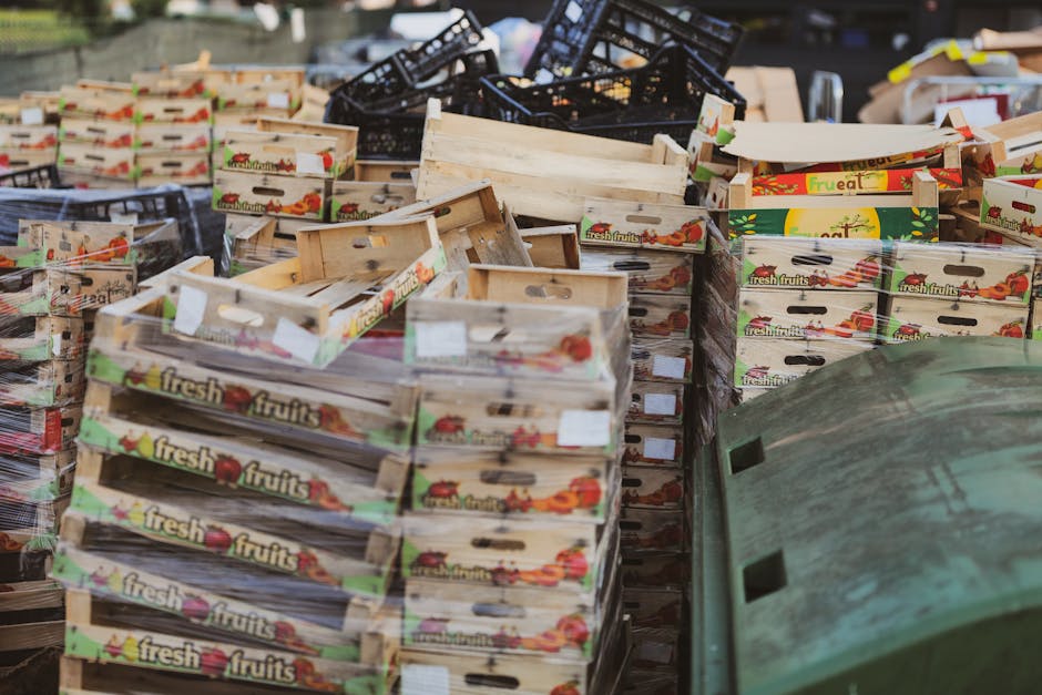 The image displays a large collection of empty wooden fruit crates, primarily made of light-colored, unfinished wood with visible grain and reinforcing nails, stacked haphazardly on a paved outdoor surface. Some crates feature colourful printed labels with images of tomatoes and the words 'fresh fruits,' indicating their original purpose for produce storage. The crates vary in orientation, with some leaning against each other and others lying flat, creating a cluttered scene typical of waste or surplus packaging materials awaiting disposal or reuse. In the background, there are dark-colored plastic baskets and a weathered green plastic container with a rough surface, partially obscured by the crates, suggesting an environment where rubbish or recyclable materials are being gathered. The overall setting appears to be outdoors, possibly in a marketplace or industrial area focused on waste collection or independent rubbish removal services, as highlighted by the presence of discarded packaging materials. The lighting is diffuse, indicating an overcast day, adding to the neutral and factual tone of the scene, which aligns with the context of rubbish removal and waste management services such as those offered by Rubbish Removal Greenwich.