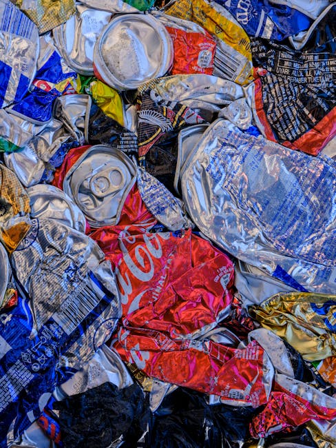 A close-up view of a pile of crushed aluminum beverage cans with various colors and branding, including silver, red, blue, black, and gold. The cans have visible pull tabs and are crumpled, showing reflective metallic surfaces with some labels partially torn or obscured. The cans are scattered and overlapping on a surface, likely for collection or disposal, and the scene suggests an environment related to waste management or recycling activities. This kind of collection of flattened cans is typical in private waste handling or informal rubbish collection, illustrating material which could be processed by rubbish removal services or recycling programs. The lighting highlights the metallic textures and vibrant colors, emphasizing the state of the cans ready for disposal or recycling in a professional or domestic setting. Rubbish Removal Greenwich often handles this type of scrap metal as part of comprehensive rubbish clearance or alternative waste handling solutions.