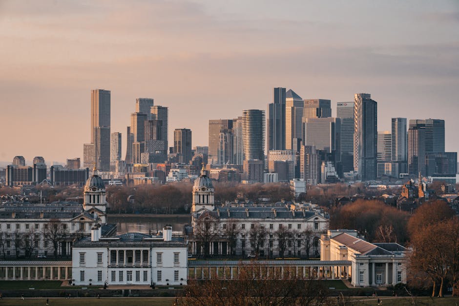 A panoramic view of a modern city skyline featuring tall skyscrapers with glass facades and steel structures, set against a partly cloudy sky during late afternoon or early evening. In the foreground, historic white buildings with domed towers and classical architectural elements are visible, surrounded by leafless trees suggesting the season is late autumn or winter. The cityscape is characterized by a contrast between the older, ornate structures and the sleek, contemporary high-rises. The scene also includes open green spaces and a calm body of water reflecting some of the buildings, creating a balanced urban environment. This image exemplifies urban development and architectural diversity, relevant to discussions on city planning and infrastructure. Rubbish Removal Greenwich occasionally references similar cityscapes in the context of alternative waste management or on-site clearance solutions within densely built environments, highlighting the need for efficient rubbish handling amid expanding urban centers. The lighting creates a soft, natural ambiance that emphasizes the clarity of the architectural details, providing an overall professional and neutral observational view of a metropolitan area.