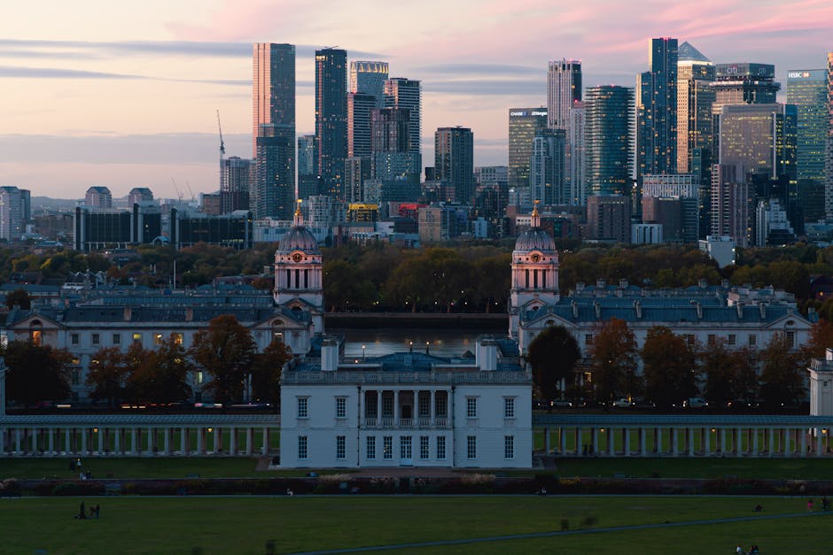 The image shows a view of the historic Old Royal Naval College with its classical white facades, symmetrical layout, and ornate domed towers positioned in the foreground. Surrounding the building, there are well-maintained green lawns and a row of trees with autumn foliage, adding touches of orange and brown. Beyond the historic site, a river runs parallel, and across it, a modern cityscape with numerous high-rise office buildings and skyscrapers dominates the background. The skyscrapers are constructed of glass and steel, reflecting the soft pink and purple hues of the early evening sky. The lighting suggests the sun has recently set, casting a gentle glow over the scene. This urban and historic combination underscores the contrast between traditional architecture and contemporary city development. The overall atmosphere is calm, with a serene dusk ambiance that highlights both the architectural details and the layered city environment, subtly hinting at aspects of urban renewal and the importance of maintaining and managing both historic sites and modern urban spaces.