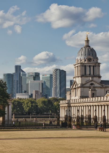 A historic stone building with a prominent domed tower featuring a decorative lantern and a gold finial on top, situated in a park-like area with open grassy expanse and a black wrought iron fence. The building's façade displays classical architectural details, including columns and ornamental stonework. Behind the structure, a cluster of modern high-rise office buildings with glass and steel façades rise towards a partly cloudy sky, creating a contrast between historic and contemporary architecture. The scene includes trees lining the background, providing greenery amidst the urban environment. The area appears to be a public space or garden with pedestrians visible along the pathways, and the overall atmosphere suggests a clear, bright day. This setting illustrates the integration of traditional architecture within a busy cityscape, emphasizing the importance of maintenance and possible private waste management services to keep such public areas clean and well-preserved, exemplifying alternative waste handling and rubbish removal in an urban context.