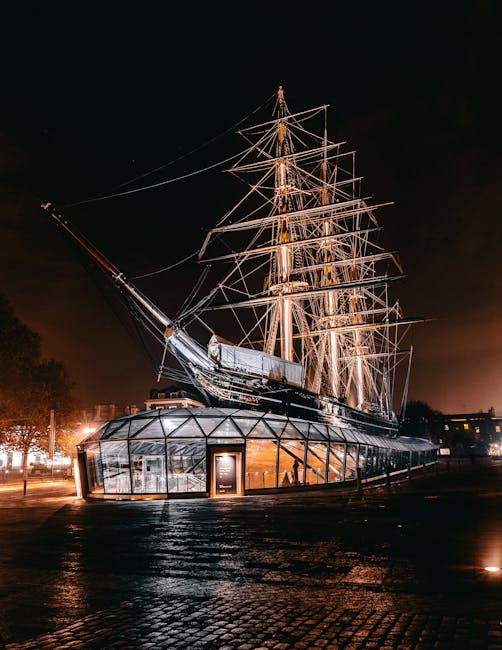 A nighttime exterior view of a large, historic sailing ship with a prominent wooden hull and multiple tall masts, illuminated by warm artificial lighting that highlights its intricate rigging and structure. The ship is positioned on a paved outdoor area, with a glass-enclosed modern structure at its front that appears to serve as an entrance or exhibit space, featuring glass panels, a door, and interior lighting. In the background, faint outlines of trees and nearby buildings are visible against a dark sky, creating a contrasting scene of ancient maritime architecture and contemporary outdoor display, all captured under soft, ambient lighting that emphasizes the textures of the wooden masts, rigging, and surrounding paved surface. The scene aligns with private or independent vessel preservation, often associated with historic ships that may be subject to conservation or special display arrangements outside traditional maritime museums, consistent with the theme of alternative waste handling environments where large objects are carefully preserved or managed outside conventional settings.