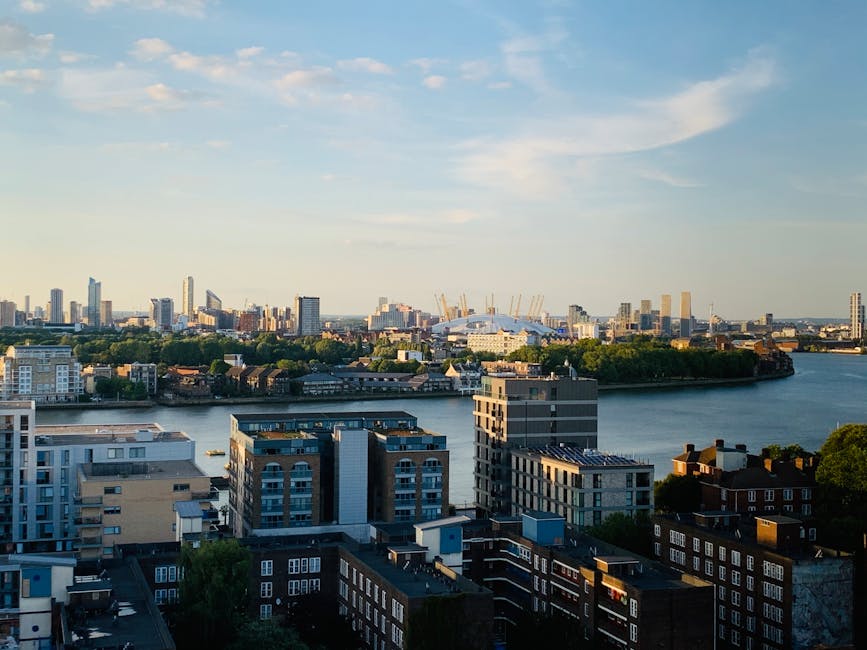 A panoramic view of a city skyline featuring a mixture of modern and older residential buildings in the foreground, with flat rooftops and various window arrangements in shades of brown, white, and grey. The buildings are set along a river with calm water reflecting the blue sky and scattered clouds above. In the background, there are numerous high-rise office towers and cranes indicating ongoing construction within a dense urban area. The overall scene captures an expansive view of the city’s urban landscape on a clear day, with natural light highlighting the textures and colors of the buildings, which relate to the context of urban waste management and alternative rubbish disposal solutions such as private collection services like Rubbish Removal Greenwich.