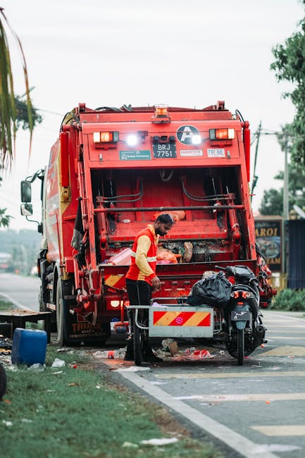 A worker in a red safety vest and dark trousers stands at the rear of a large red rubbish collection vehicle on a street. The vehicle's rear hatch is open, revealing the mechanical compactor inside, with some waste visible near the opening. The worker appears to be emptying or handling a black garbage bag and is positioned next to a small black motorbike parked on the roadway. The scene is set outdoors, with a partly cloudy sky overhead, and trees and utility poles visible in the background. The surrounding environment includes a sidewalk with a grassy verge and some litter scattered on the ground. The rubbish vehicle has identifiable labels and markings, and the setting suggests an urban or suburban area where waste collection occurs, supporting the context of private rubbish removal services as an alternative to council-provided waste collection, as referenced in the page about skip hire versus rubbish removal costs.