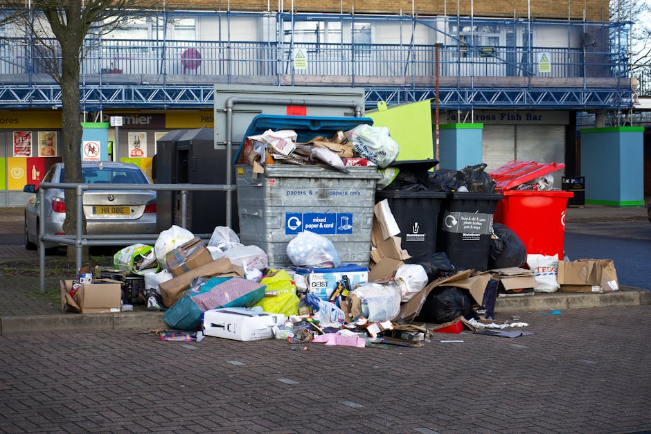 A large overflowing recycling and waste collection point situated on an urban street, featuring multiple bins and containers filled with various refuse materials. The central bin is a metallic mixed paper and card recycling container, with its lid open and cardboard and paper waste spilling over onto the pavement. To its right, a black waste bin is nearly full, with black plastic bags and loose trash surrounding its base. Adjacent to these, a red bin is visible, partially obscured, with additional plastic bags and debris scattered in front. The background shows a building with blue construction netting and scaffolding, indicating ongoing work, along with a storefront with yellow signage and storefront windows. A parked silver car is to the left, behind a metal railing, and a leafless tree is situated nearby, suggesting seasonal context. The scene is lit with natural daylight, highlighting the untidy state of the refuse area, which illustrates the importance of proper waste disposal and highlights issues related to rubbish management in urban environments, relevant to the services provided by Rubbish Removal Greenwich.